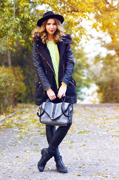 Fashion portrait of pretty young blonde smiling woman wearing trendy coat, vintage hat and neon sweater, posing at countryside park in nice sunny fall autumn day.