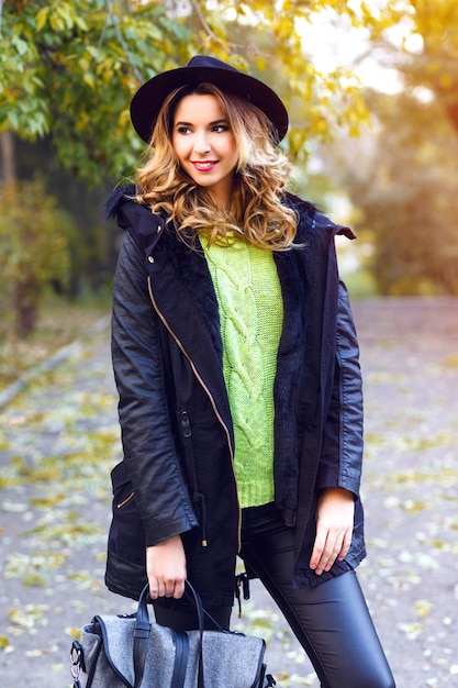 Fashion portrait of pretty young blonde smiling woman wearing trendy coat, vintage hat and neon sweater, posing at countryside park in nice sunny fall autumn day.