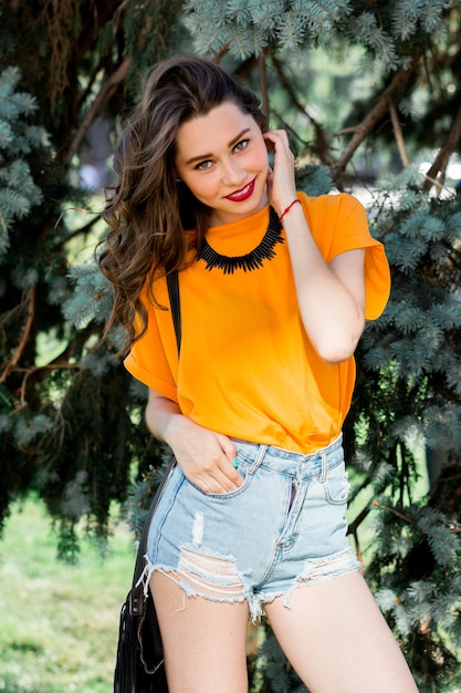 Fashion portrait of pretty young woman in the summer park wearing orange shirt and stylish jeans shorts with boho bag.