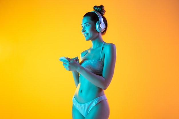 Fashion portrait of seductive girl in stylish swimwear on yellow wall in neon. Summertime, beach season. Listen to music with phone and headphones, dancing. Resort, vacation, holidays concept.