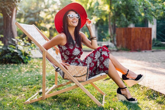 유행 액세서리, 지갑, 선글라스, 빨간 모자를 쓰고 여름 옷을 입고 포즈를 취하는 매력적인 세련된 여성 미소의 패션 초상화, 갑판 의자에서 휴가 휴식