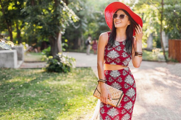 Модный портрет улыбающейся привлекательной стильной женщины, идущей в парке в летнем наряде с принтом, в модных аксессуарах, сумочке, солнцезащитных очках, красной шляпе, отдыхающей в отпуске