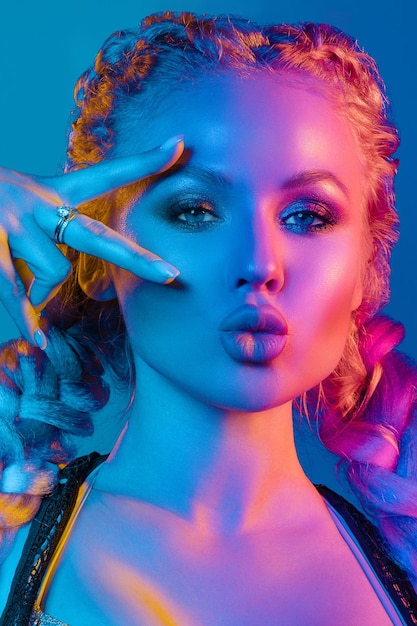 Fashion portrait of stunning sexy young blonde woman with trendy braids and sparkling bra under the net top she is holding peace sign by the eye making pouting lips in bright blue and red light