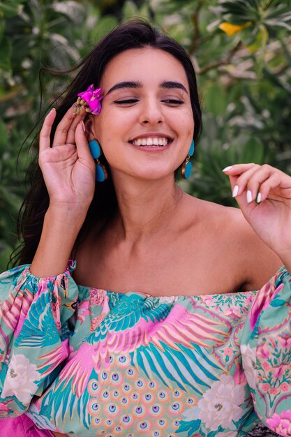 Fashion portrait of stylish woman in colorful print long sleeve top and pink shorts on beach, tropical background.