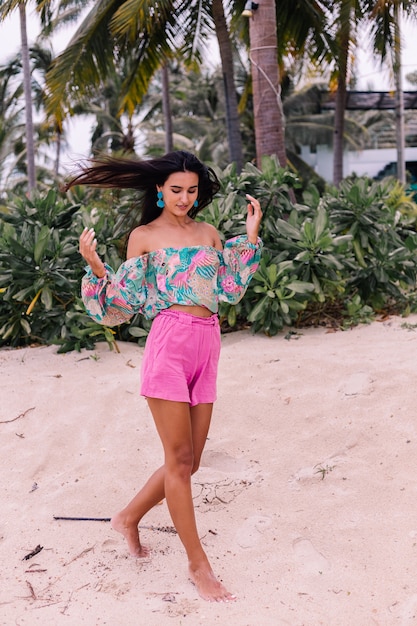 Fashion portrait of stylish woman in colorful print long sleeve top and pink shorts on beach, tropical background.