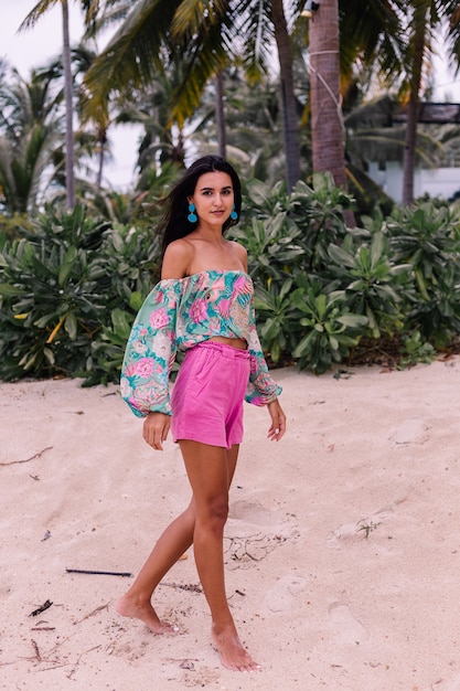 カラフルなプリントの長袖トップとビーチ、熱帯の背景にピンクのショートパンツでスタイリッシュな女性のファッションの肖像画。