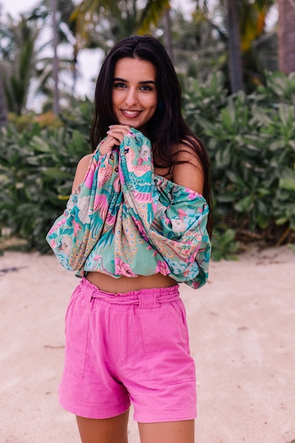 Fashion portrait of stylish woman in colorful print long sleeve top and pink shorts on beach, tropical background.