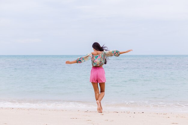 Fashion portrait of stylish woman in colorful print long sleeve top and pink shorts on beach, tropical background.