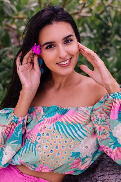 Fashion portrait of stylish woman in colorful print long sleeve top and pink shorts on beach, tropical background.