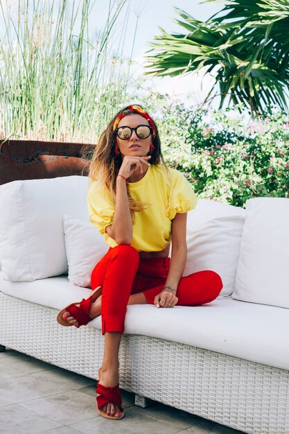 Фасонируйте портрет стильной женщины в красных штанах, желтом топе, солнечных очках и головном платке.