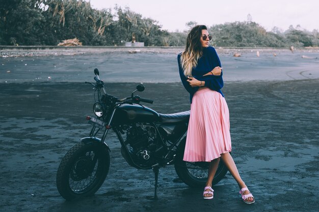 Fashion portrait of stylish woman in sunglasses, blue sweater and pink skirt, stands by black bike on black sand beach.