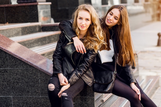 Fashion portrait of Two stylish pretty women posing on the street in sunny day. Wearing trendy urban outfit , leather jacket and boots heels. Young friends  waiting on stairs outdoor.