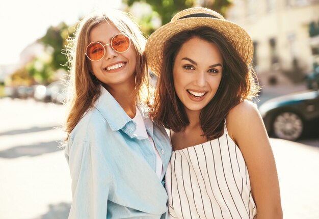 Fashion portrait of two young smiling stylish hippie brunette and blond women models in summer sunny day in hipster clothes posing on the street background