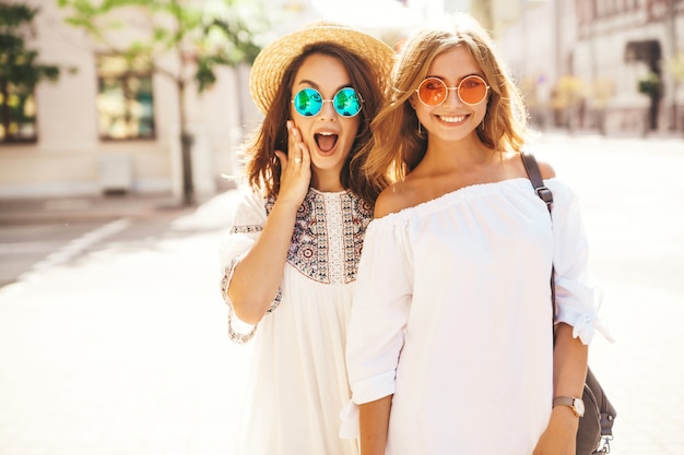 Fashion portrait of two young stylish hippie brunette and blond women models. Best friends in white summer hipster dress posing