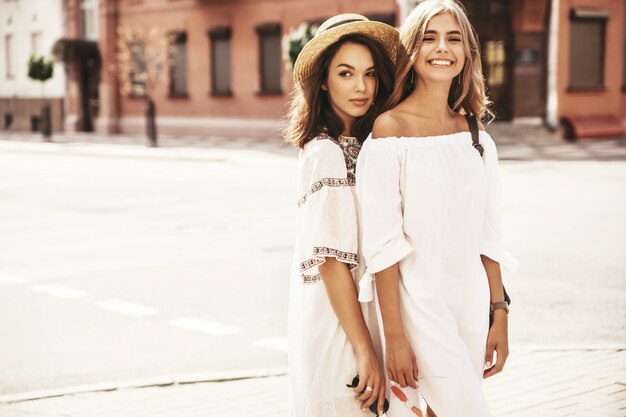 Fashion portrait of two young stylish hippie brunette and blond women models in summer sunny day in white hipster clothes posing . No makeup