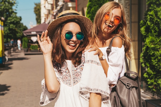 Fashion portrait of two young stylish hippie brunette and blond women models in summer sunny day in white hipster clothes posing