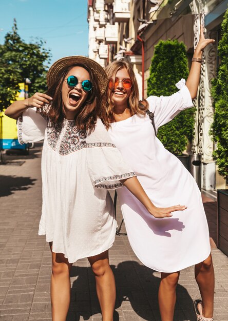 Fashion portrait of two young stylish hippie brunette and blond women models in summer sunny day in white hipster clothes posing