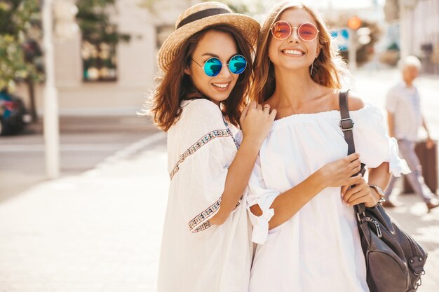 Fashion portrait of two young stylish hippie brunette and blond women models in summer sunny day in white hipster clothes posing