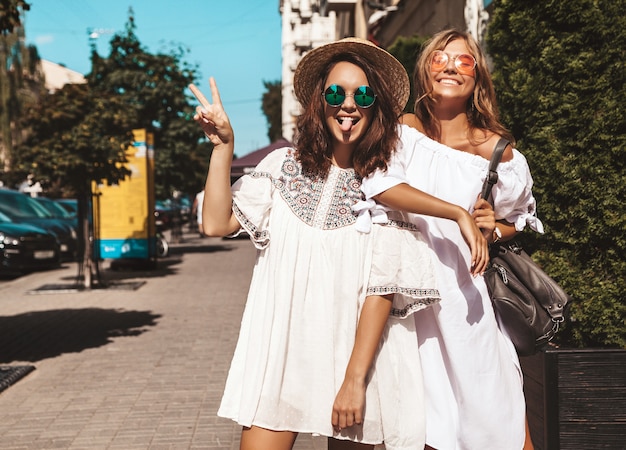 Fashion portrait of two young stylish hippie brunette and blond women in summer sunny day. Models dressed in white hipster clothes. womans posing