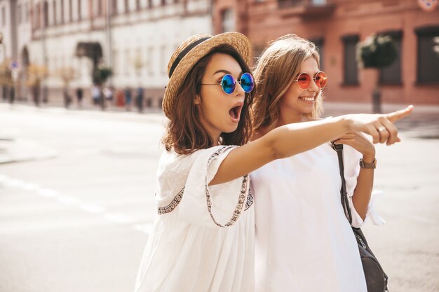 Adatti un ritratto di due donne castane e bionde di giovane giovane hippy alla moda nel giorno soleggiato dell'estate nella posa bianca dei vestiti dei pantaloni a vita bassa