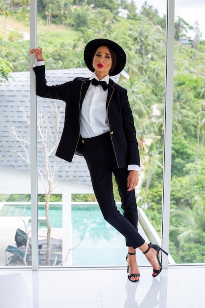 Fashion portrait of woman in black suit, butterfly tie and hat at luxury villa