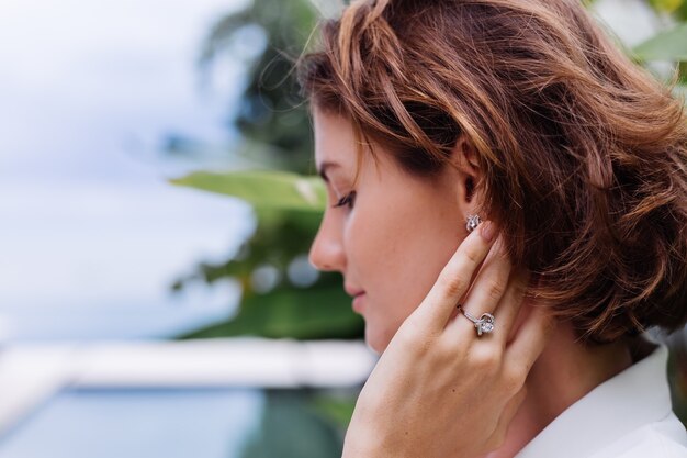 Fashion portrait of woman at tropical luxury villa wearing white stylish blazer and jewellery over tropical leaves