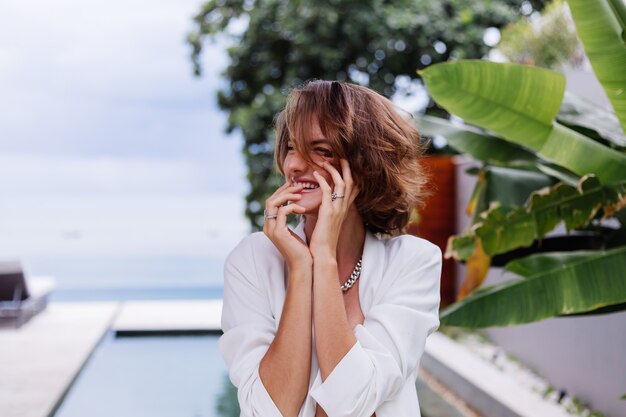 Fashion portrait of woman at tropical luxury villa wearing white stylish blazer and jewellery over tropical leaves