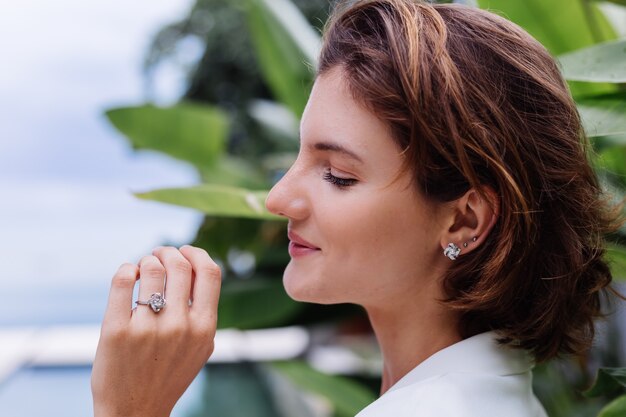 Fashion portrait of woman at tropical luxury villa wearing white stylish blazer and jewellery over tropical leaves