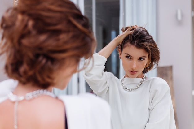 Fashion portrait of young caucasian woman professional model in white blazer and silver chain look in mirror at luxury villa