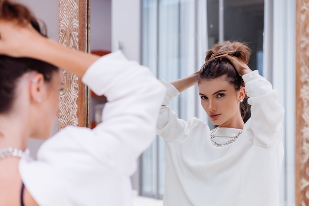 Фасонный портрет молодой кавказской профессиональной модели в белом пиджаке и серебряной цепочке смотрится в зеркало на роскошной вилле