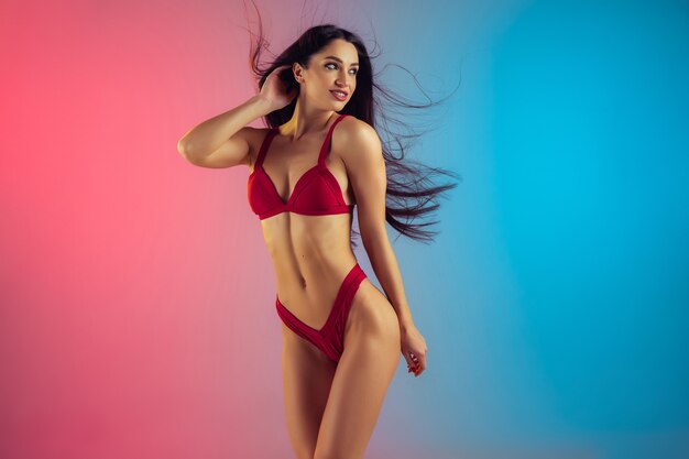 Модный портрет молодой подтянутой и спортивной женщины в стильных красных роскошных купальниках на градиентной стене с идеальным телом, готовым к лету