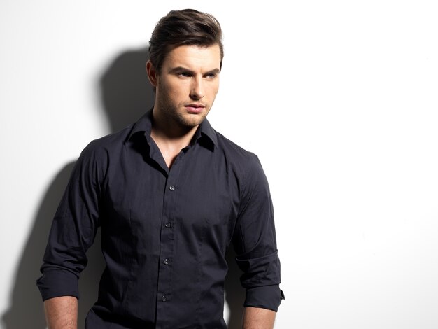 Fashion portrait of young man in black shirt poses over wall with contrast shadows