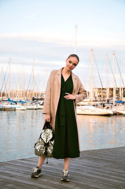 Free photo fashion portrait of young stunning elegant woman posing  promenade, wearing dress coat sneakers and backpack, luxury tourist, soft warm colors.