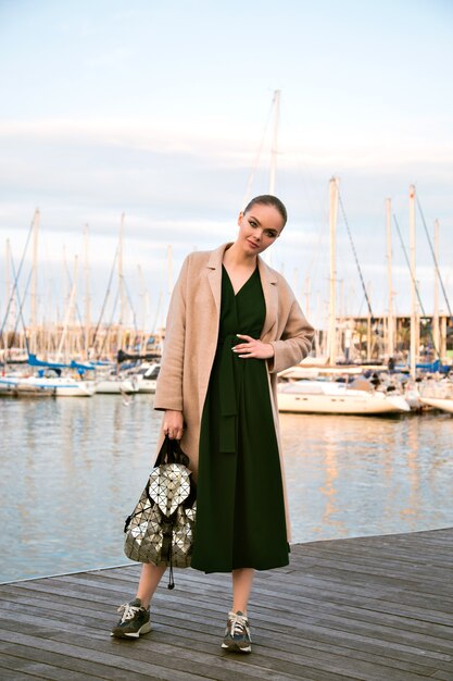 Fashion portrait of young stunning elegant woman posing  promenade, wearing dress coat sneakers and backpack, luxury tourist, soft warm colors.