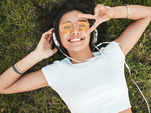 Fashion portrait of young stylish hipster woman lying on the grass in the park .Girl weares trendy outfit.Smiling model enjoy her weekends. Female listening to music via headphones.Top view