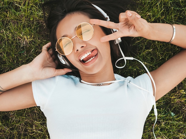 Fashion portrait of young stylish hipster woman lying on the grass in the park .Girl weares trendy outfit.Smiling model enjoy her weekends. Female listening to music via headphones.Top view