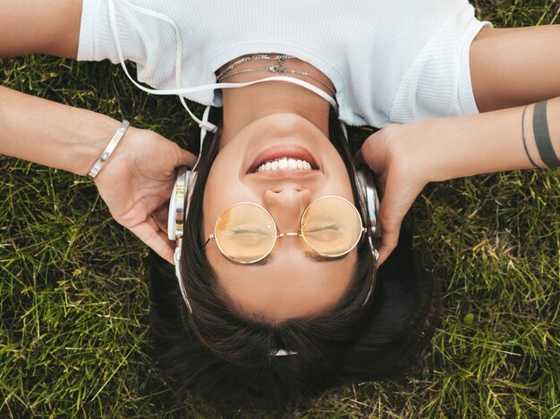 Fashion portrait of young stylish hipster woman lying on the grass in the park .Girl weares trendy outfit.Smiling model enjoy her weekends. Female listening to music via headphones.Top view