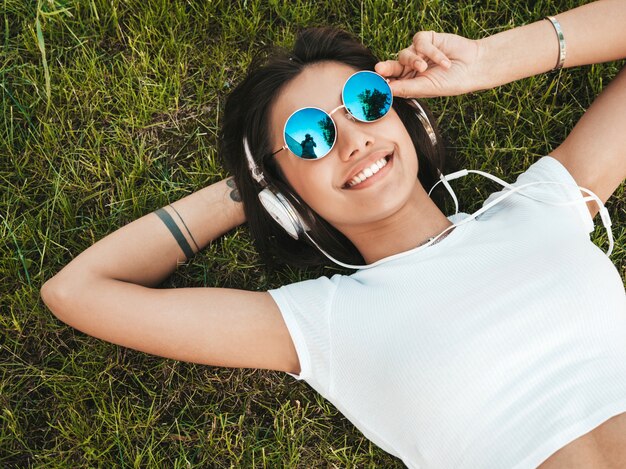 Fashion portrait of young stylish hipster woman lying on the grass in the park .Girl weares trendy outfit.Smiling model enjoy her weekends. Female listening to music via headphones.Top view