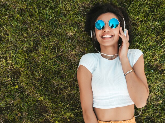 Fashion portrait of young stylish hipster woman lying on the grass in the park .Girl weares trendy outfit.Smiling model enjoy her weekends. Female listening to music via headphones.Top view