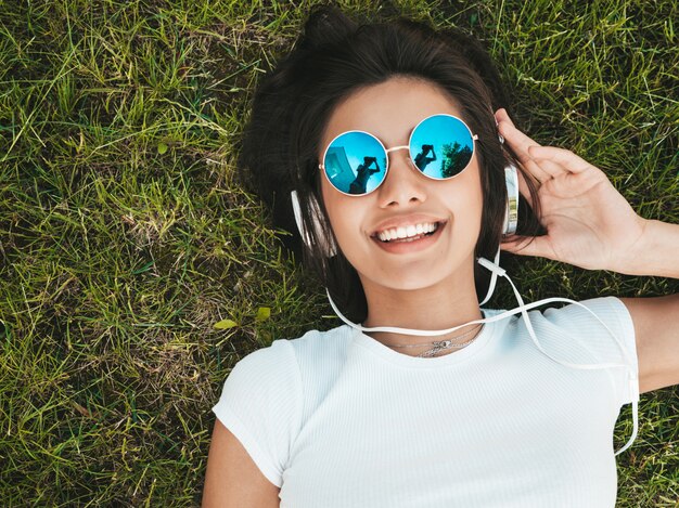 Fashion portrait of young stylish hipster woman lying on the grass in the park .Girl weares trendy outfit.Smiling model enjoy her weekends. Female listening to music via headphones.Top view