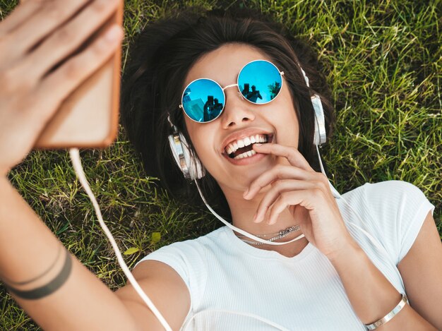 Fashion portrait of young stylish hipster woman lying on the grass in the park .Girl weares trendy outfit.Smiling model making selfie. Female listening to music via headphones.Top view