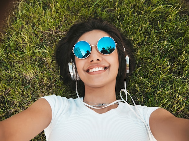 Fashion portrait of young stylish hipster woman lying on the grass in the park .Girl weares trendy outfit.Smiling model making selfie. Female listening to music via headphones.Top view