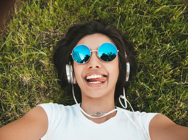 Fashion portrait of young stylish hipster woman lying on the grass in the park .girl weares trendy outfit.smiling model making selfie. female listening to music via headphones.top view