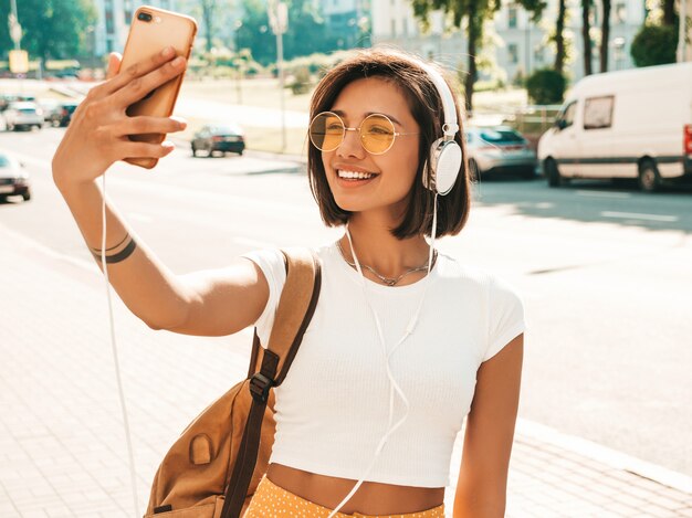 通りを歩いている若いスタイリッシュな流行に敏感な女性のファッションの肖像画。女の子はselfieを作る。笑顔のモデルは、バックパックで彼女の週末をお楽しみください。ヘッドフォンで音楽を聴く女性