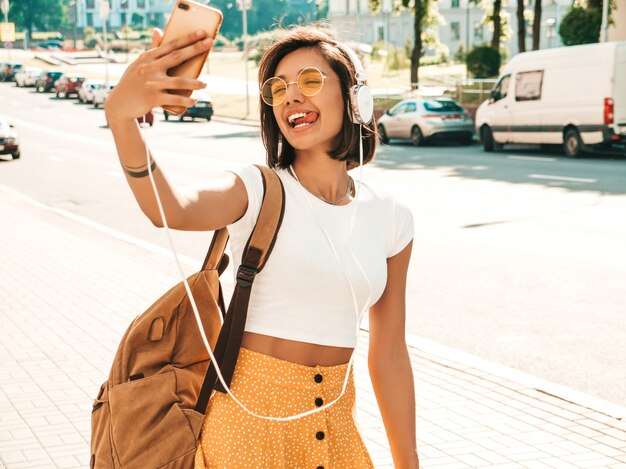 通りを歩いている若いスタイリッシュな流行に敏感な女性のファッションの肖像画。女の子はselfieを作る。笑顔のモデルは、バックパックで彼女の週末をお楽しみください。ヘッドフォンで音楽を聴く女性