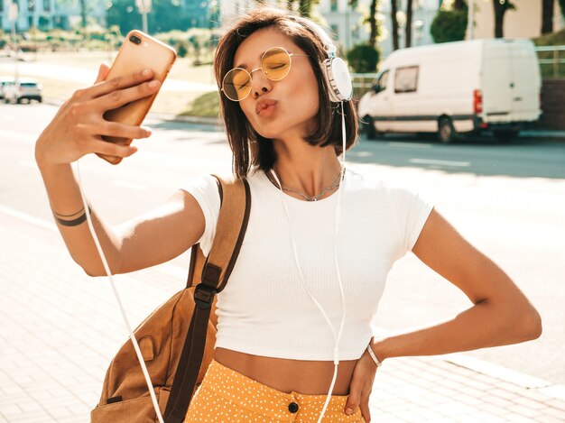 Fashion portrait of young stylish hipster woman walking in the street.Girl making selfie.Smiling model enjoy her weekends with backpack. Female listening to music via headphones