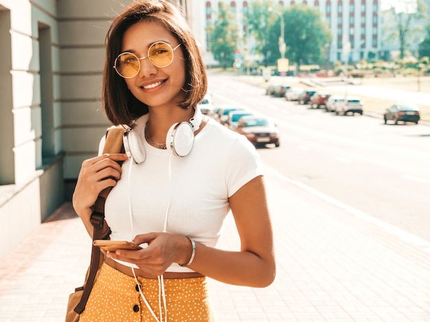 通りを歩いている若いスタイリッシュな流行に敏感な女性のファッションの肖像画。かわいいトレンディな服を着ている女の子。笑顔のモデルは彼女の週末を楽しんで、バックパックで旅行します。ヘッドフォンで音楽を聴く女性