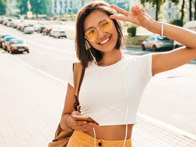 Fashion portrait of young stylish hipster woman walking in the street.Girl wearing cute trendy outfit.Smiling model enjoy her weekends, travel with backpack. Female listening to music via headphones