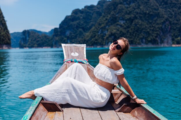 Fashion portrait of young woman in white top and pants on vacation, on sailing thai wooden boat. Travel concept. Female in Khao Sok National park.