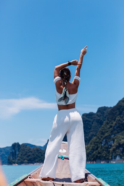 Fashion portrait of young woman in white top and pants on vacation, on sailing thai wooden boat. Travel concept. Female in Khao Sok National park.